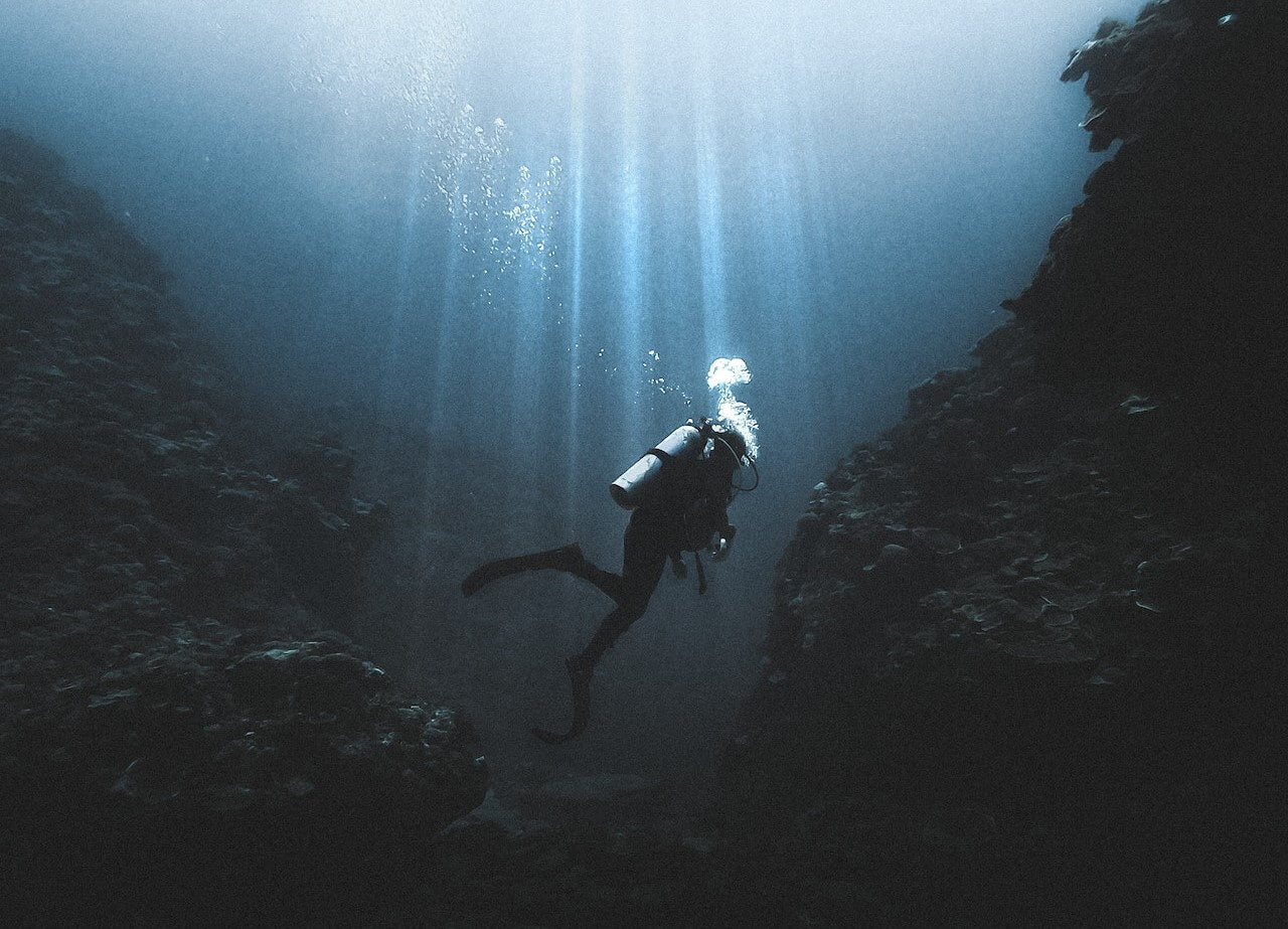 A scuba diver exploring a dark underwater section with sunlight streaming from above.